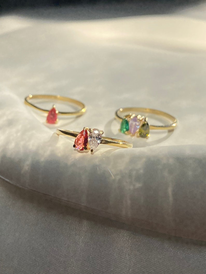 May include: Three gold rings with pear-shaped gemstones. The first ring has a red gemstone, the second ring has a clear gemstone, and the third ring has three gemstones: green, purple, and green.