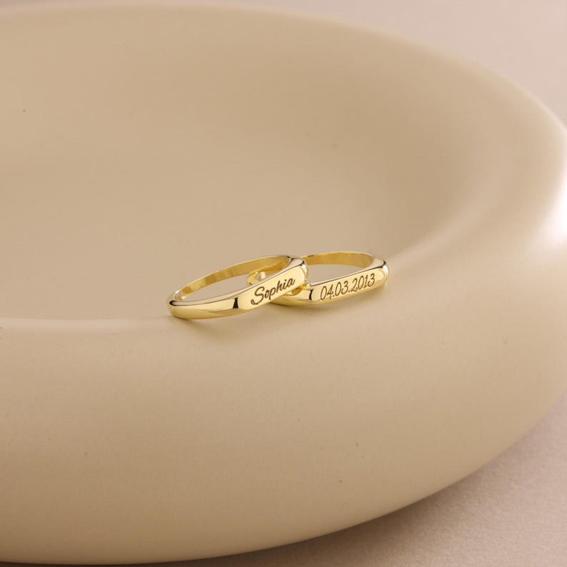 May include: Two gold rings with engraved text. One ring is inscribed with the name "Sophia", and the other with the date "04.03.2013". The rings are displayed on a light beige surface.