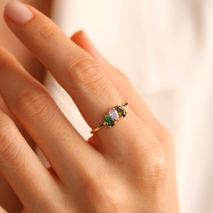 May include: A gold ring with three pear-shaped gemstones. The gemstones are green, green, and purple.