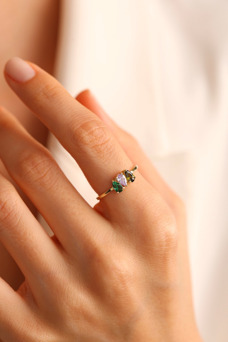 May include: A gold ring with three pear-shaped gemstones. The gemstones are green, green, and purple.