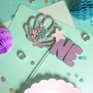 May include: A pastel-colored cake topper with a seashell, seahorse, and starfish design. The word "ONE" is displayed in purple letters. The topper is on a clear stick, with confetti and decorative paper elements.