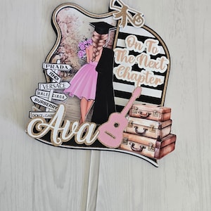 May include: A graduation cake topper featuring a cartoon illustration of a graduate in a pink dress and black gown. The topper includes the name "Ava", the phrase "On To The Next Chapter", a pink guitar, and stacked suitcases. Fashion brand signs are also included.