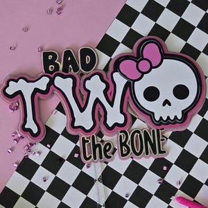 May include: A pink and black cake topper with the words "BAD TWO the BONE" and a skull with a pink bow. The background includes a pink surface and a black and white checkered pattern.