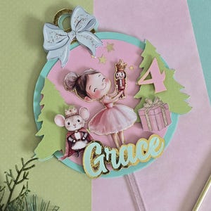 May include: A whimsical birthday-themed ornament featuring a ballerina, a mouse, and a nutcracker. The design includes the number 4, a gift, and the name "Grace" in a decorative font. The color palette is soft pink, green, and gold.