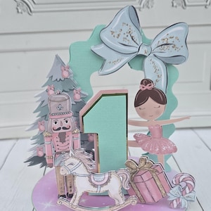 May include: A decorative centerpiece featuring a large number one in mint green, accented by a nutcracker, a ballerina, a rocking horse, a gift box, a candy cane, and a Christmas tree, all topped with a large light blue bow on a round base.