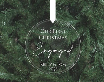 May include: Clear round Christmas ornament with the text "Our First Christmas Engaged Kelly & Tom 2023" on a green Christmas tree.