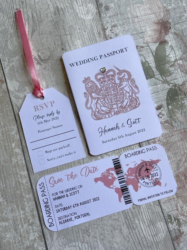 May include: A white wedding invitation set with a pink ribbon. The invitation features a passport design with a royal crest and the text "Wedding Passport". The RSVP card has a luggage tag design with the text "RSVP" and "Please reply by 6th May 2022". The save the date card has a boarding pass design with the text "Save the Date" and "Hannah & Scott".