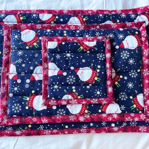 May include: Set of four quilted pot holders featuring a festive Christmas pattern with Santa Claus and snowflakes on a dark blue background. The pot holders have a red border with a white snowflake pattern.