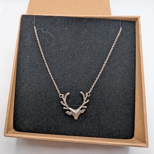 May include: A silver-toned metal necklace featuring a pendant shaped like a deer head with prominent antlers. The delicate chain connects to the sides of the antlers. The necklace is presented inside a square cardboard box with a black foam insert.