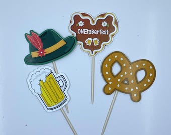 May include: Four decorative cupcake toppers featuring Oktoberfest themes: a green Tyrolean hat with a red feather, a yellow beer mug with white foam, a brown heart-shaped gingerbread cookie with the text ONEtoberfest, and a golden brown pretzel.