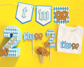 May include: A party decoration set featuring a white t-shirt with the text "two" and a pretzel graphic, a "two" banner, a large 3D number "2" with the name "Antonio", and several small pretzel-themed cupcake toppers in blue and white.