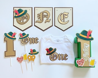May include: A set of Oktoberfest-themed party decorations, including a banner with the word "ONE" in gold letters, a green and gold number one with a felt hat, and a white onesie with a gold "ONE" and a felt hat.
