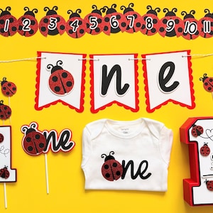 May include: A collection of ladybug-themed party decorations on a yellow background. Includes a banner with ladybug accents and numbers, a "one" banner, cake toppers, a white onesie, and a large number "1" with ladybug details. The decorations are red, black, and white.