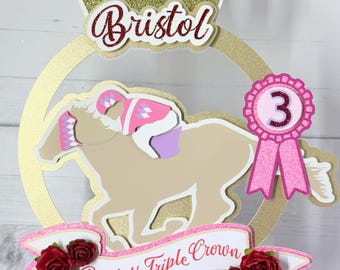 May include: A gold and pink cake topper with a horse, a crown, a ribbon with the number 3, and a banner that says "Bristol's Triple Crown".