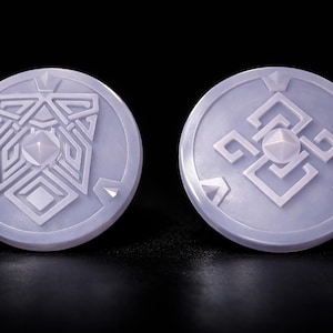 May include: Two circular, pale purple pendants with detailed geometric patterns. Each pendant has a central, raised, diamond-shaped feature. The pendants are displayed against a black backdrop, emphasising their intricate design.