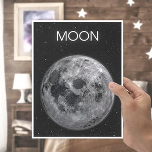 May include: A print featuring a detailed image of the moon against a starry background. The word "MOON" is displayed in white at the top. The print is held by a hand, with a wooden wall and decorative stars in the background.