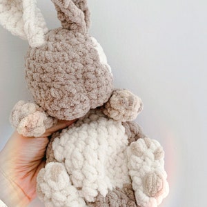 Puede incluir: Un juguete de peluche de conejo tejido a crochet con pelo marr&oacute;n y blanco. El conejo est&aacute; sentado con las orejas levantadas y las patas metidas.