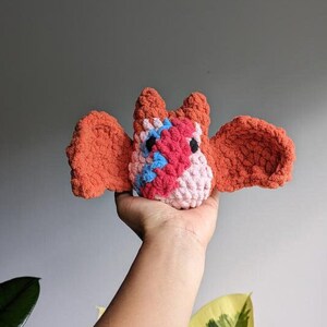 May include: A crocheted bat plush toy with orange wings and a pink, red, and blue body. The toy is being held in a person's hand.