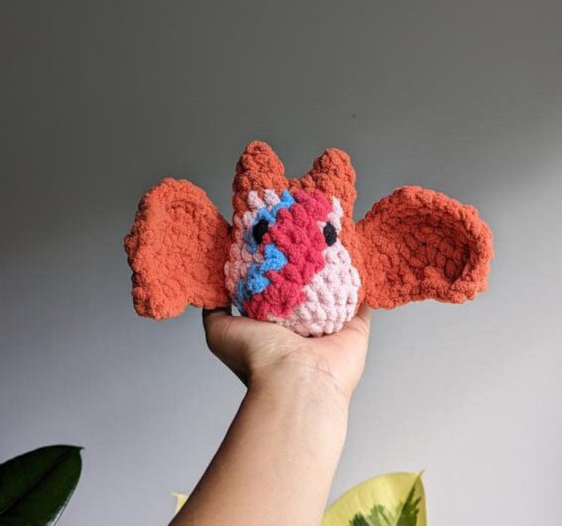 May include: A crocheted bat plush toy with orange wings and a pink, red, and blue body. The toy is being held in a person's hand.