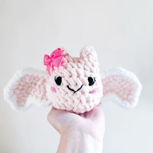 May include: A pink crocheted plush toy with white wings and a pink bow. The toy has a smiling face with black eyes and a pink nose.