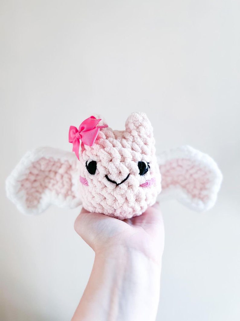 May include: A pink crocheted plush toy with white wings and a pink bow. The toy has a smiling face with black eyes and a pink nose.