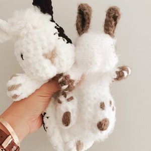 Puede incluir: Dos animales de peluche tejidos a crochet, uno blanco con detalles marrones y el otro blanco con detalles negros, se sostienen en una mano. El animal de peluche blanco con detalles marrones tiene orejas largas.