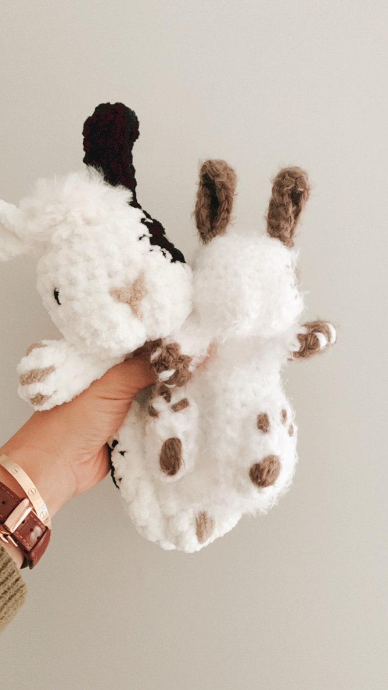 Puede incluir: Dos animales de peluche tejidos a crochet, uno blanco con detalles marrones y el otro blanco con detalles negros, se sostienen en una mano. El animal de peluche blanco con detalles marrones tiene orejas largas.