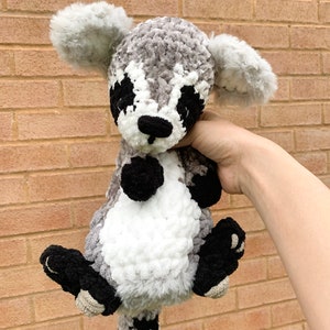 May include: A crocheted raccoon plush toy with gray, white, and black fur. The toy has a black nose and eyes, and a white stripe on its face.