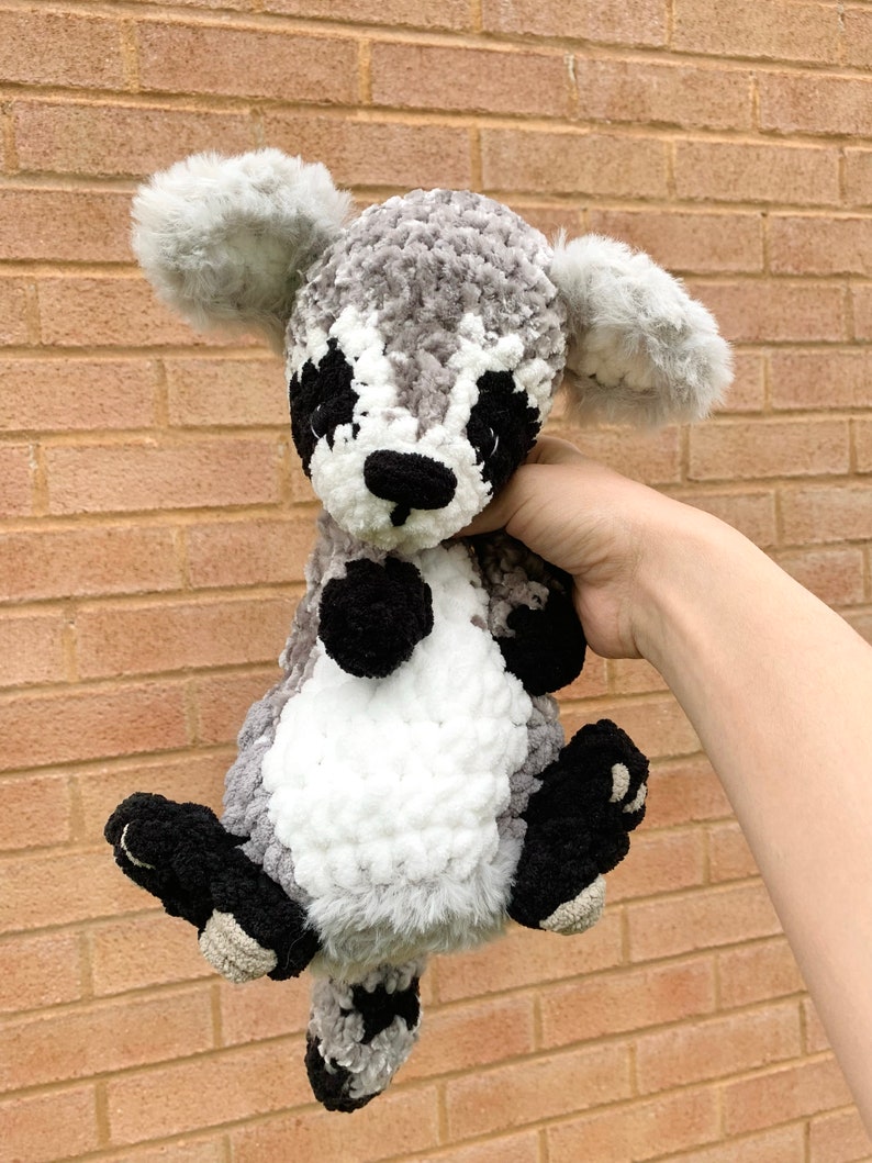 May include: A crocheted raccoon plush toy with gray, white, and black fur. The toy has a black nose and eyes, and a white stripe on its face.