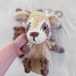 May include: A crocheted stuffed animal resembling a fawn with brown and white fur. The fawn has large ears, a brown nose, and black eyes. The fawn is being held by a hand.