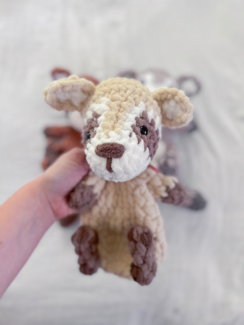 May include: A crocheted stuffed animal resembling a fawn with brown and white fur. The fawn has large ears, a brown nose, and black eyes. The fawn is being held by a hand.