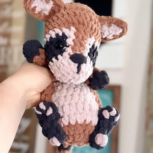 May include: A crocheted plush toy raccoon with brown, black, and pink fur. The toy has black eyes and a black nose.
