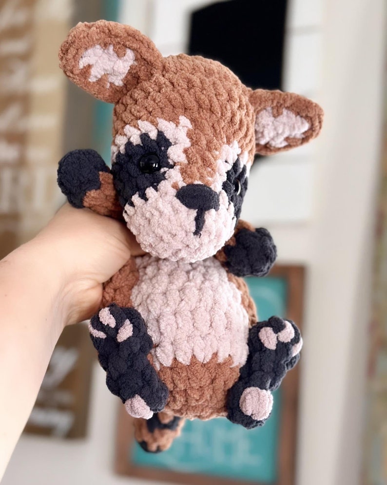 May include: A crocheted plush toy raccoon with brown, black, and pink fur. The toy has black eyes and a black nose.