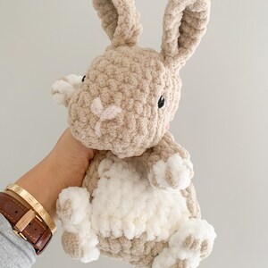 Puede incluir: Un juguete de peluche de conejo tejido a crochet con pelo marr&oacute;n y blanco. El conejo tiene orejas largas y una barriga blanca.