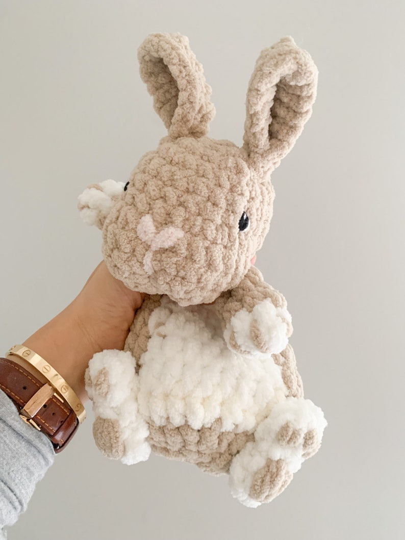 Puede incluir: Un juguete de peluche de conejo tejido a crochet con pelo marr&oacute;n y blanco. El conejo tiene orejas largas y una barriga blanca.