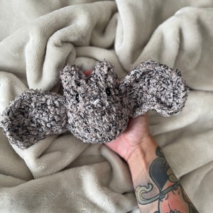 May include: A grey crocheted bat plush toy with black eyes. The toy is being held by a hand with a tattoo.