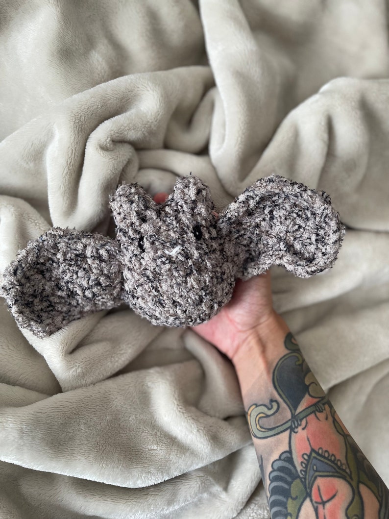 May include: A grey crocheted bat plush toy with black eyes. The toy is being held by a hand with a tattoo.
