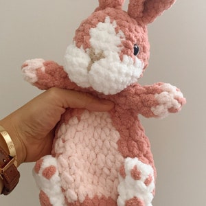 Puede incluir: Un juguete de peluche de conejo de crochet rosa y blanco con una cara blanca y ojos negros. El conejo est&aacute; sentado en su trasero con las patas extendidas.