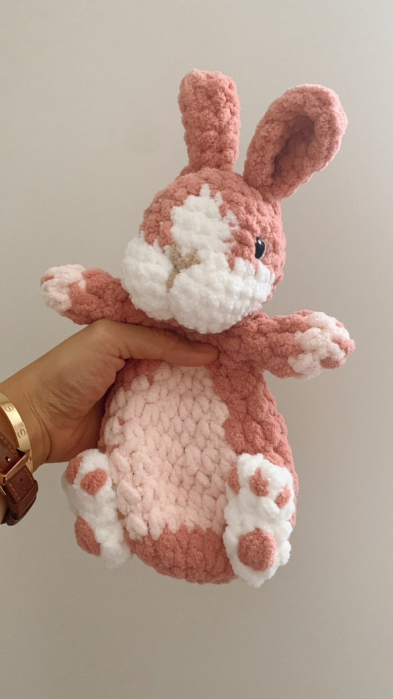 Puede incluir: Un juguete de peluche de conejo de crochet rosa y blanco con una cara blanca y ojos negros. El conejo est&aacute; sentado en su trasero con las patas extendidas.