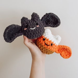 May include: A hand holds two crocheted Halloween toys. One is a black bat with a smiling face and the other is an orange, white and brown candy corn with a smiling face.