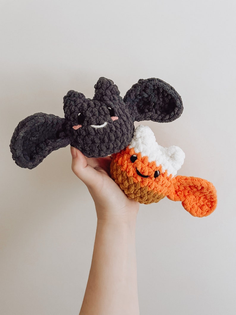 May include: A hand holds two crocheted Halloween toys. One is a black bat with a smiling face and the other is an orange, white and brown candy corn with a smiling face.