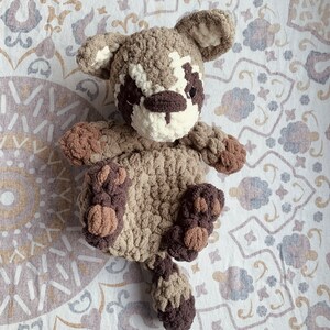May include: A crocheted stuffed animal resembling a raccoon with brown and white fur. The animal is lying on a white and brown patterned fabric.