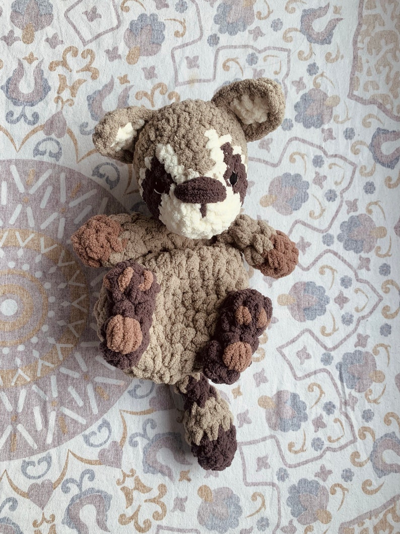 May include: A crocheted stuffed animal resembling a raccoon with brown and white fur. The animal is lying on a white and brown patterned fabric.