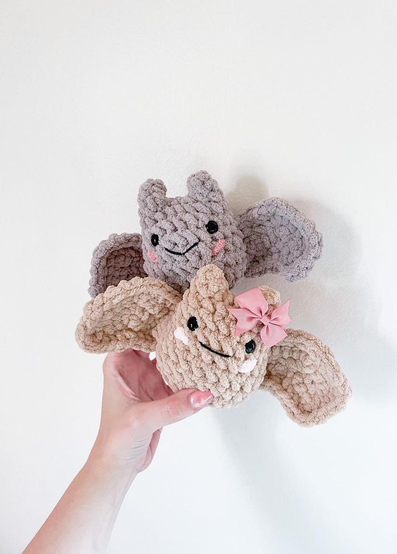 May include: Two crocheted bat plushies, one brown with a pink bow and one grey. Both have embroidered eyes and smiles.