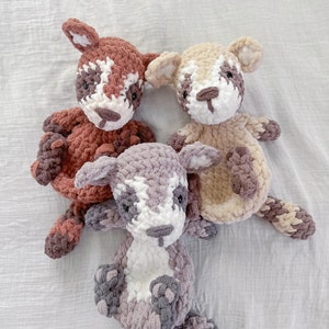 May include: Three crocheted stuffed animals in the shape of foxes. The foxes are brown, beige, and gray with white accents. The foxes are sitting on a white fabric background.