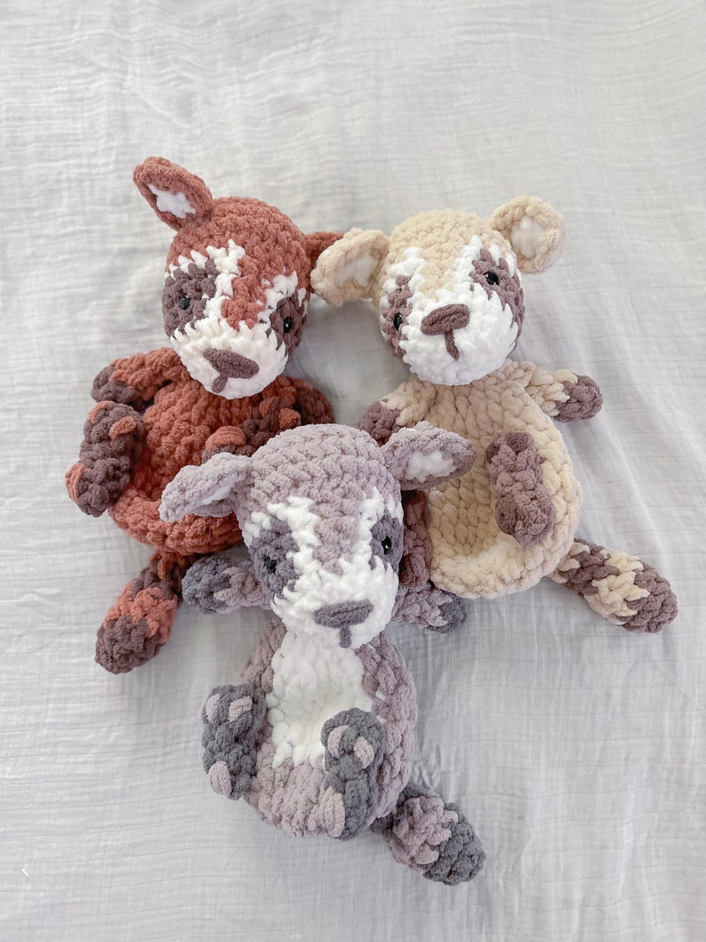 May include: Three crocheted stuffed animals in the shape of foxes. The foxes are brown, beige, and gray with white accents. The foxes are sitting on a white fabric background.