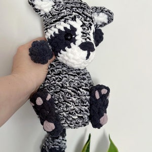May include: A crocheted raccoon plush toy with black and white fur, black eyes, and pink paw pads. The toy is being held by a hand.