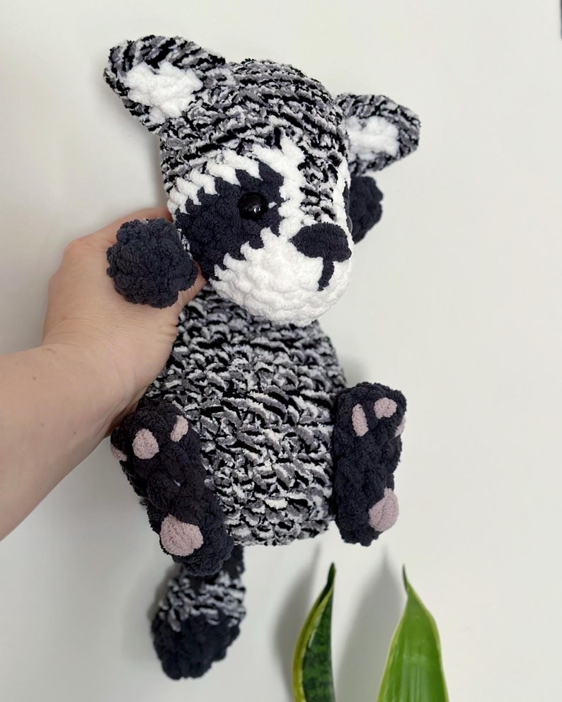 May include: A crocheted raccoon plush toy with black and white fur, black eyes, and pink paw pads. The toy is being held by a hand.
