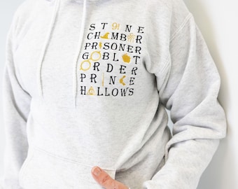 May include: A light gray hoodie with a front pocket and a hood. The hoodie features embroidered text and symbols in black and gold, including the words "Stone", "Chamber", "Prisoner", "Goblet", "Order", "Prince", and "Hallows".