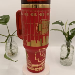 Puede incluir: Un vaso t&eacute;rmico rojo con un dise&ntilde;o dorado que presenta un campo de f&uacute;tbol americano, el logotipo de los Kansas City Chiefs y el texto "SUNDAYS FOR THE CHIEFS". El vaso tiene un asa y una tapa roja.